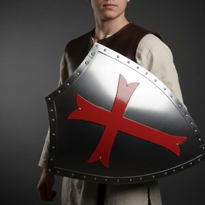 May include: A heater-style shield made of polished silver-toned metal featuring a prominent red cross emblem in the center. The shield is bordered by a metallic rim secured with evenly spaced rivets along the entire outer edge of the surface.