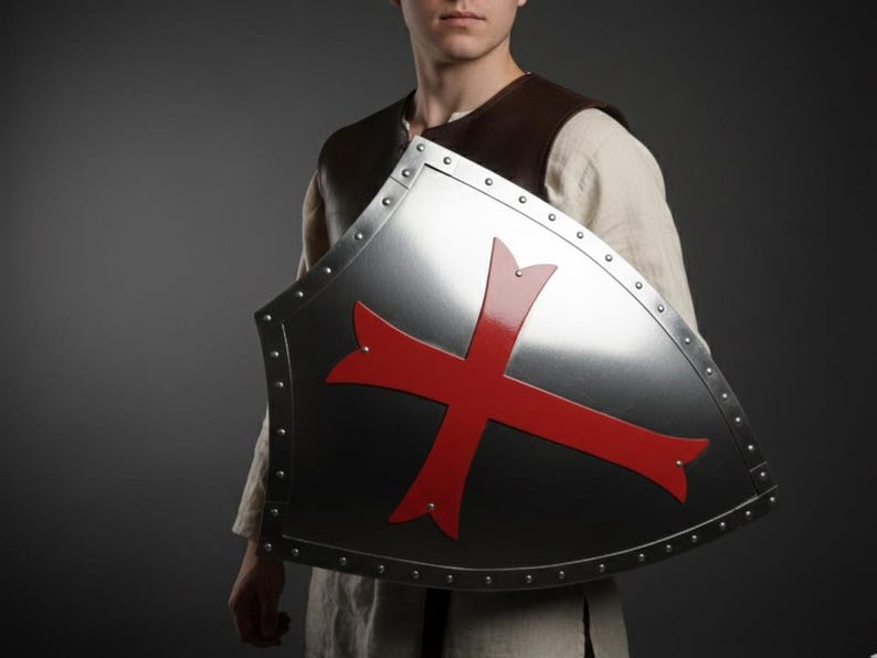 May include: A heater-style shield made of polished silver-toned metal featuring a prominent red cross emblem in the center. The shield is bordered by a metallic rim secured with evenly spaced rivets along the entire outer edge of the surface.