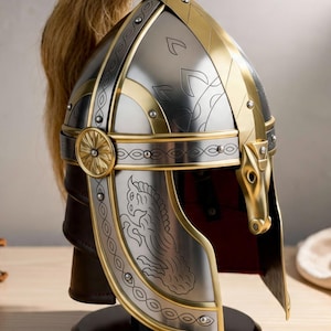 May include: A silver and gold-toned helmet with intricate detailing, including a horse head design on the side. The helmet features a blonde ponytail and is displayed on a black stand, showcasing its historical or fantasy theme.