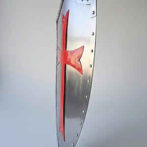 May include: A side-profile view of a metallic heater-style shield featuring a polished silver finish and a prominent red cross emblem. The shield is constructed with a reinforced border secured by a series of evenly spaced metal rivets.