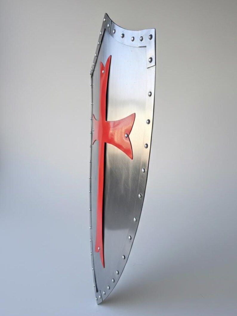May include: A side-profile view of a metallic heater-style shield featuring a polished silver finish and a prominent red cross emblem. The shield is constructed with a reinforced border secured by a series of evenly spaced metal rivets.