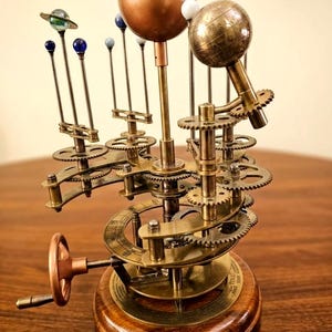 May include: A mechanical orrery model featuring a large central copper-colored sphere, a smaller etched brass globe, and several smaller colored spheres representing planets on metal arms. The device includes brass gears, a hand crank, and a wooden base.