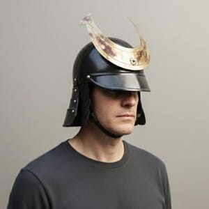 Pode incluir: Um capacete de samurai preto com viseira e crista decorativa dourada. O capacete tem abas laterais e uma correia de queixo. A pessoa na imagem está vestindo uma camiseta preta.