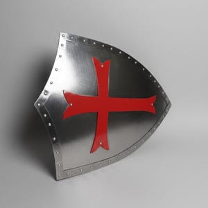 May include: A heater-style shield made of polished silver-toned metal featuring a prominent red cross emblem centered on the front surface. The shield is bordered by a metallic rim secured with evenly spaced rivets along the entire outer edge.