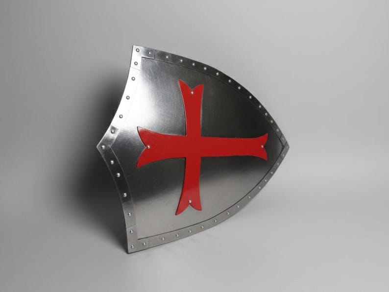 May include: A heater-style shield made of polished silver-toned metal featuring a prominent red cross emblem centered on the front surface. The shield is bordered by a metallic rim secured with evenly spaced rivets along the entire outer edge.