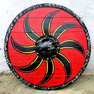 May include: A circular wooden shield featuring a vibrant red background with a black and yellow curved blade pattern radiating from the center. The shield has a black central boss and a black outer rim decorated with silver-colored metal rivets.