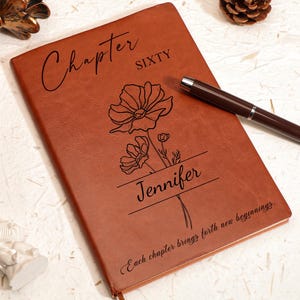 May include: A brown leather journal with the words "Chapter Sixty" and "Jennifer" engraved on the cover, along with a floral design. A pen rests on the journal. The text "Each chapter brings forth new beginnings" is also engraved.