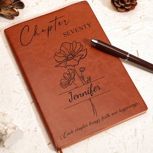 May include: A brown leather journal with the words "Chapter Seventy" and "Jennifer" engraved on the cover. The journal features a floral design and the phrase "Each chapter brings forth new beginnings."
