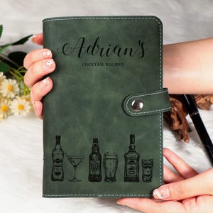 May include: A dark green leatherette cocktail recipe book with the name "Adrian's" in script. The cover features etched illustrations of cocktail glasses and bottles. The book is held open by a silver snap closure.
