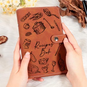 May include: A brown leather recipe book with a snap closure, featuring engraved cooking-themed illustrations. The cover reads "Recipes Book" in script. A black pen rests nearby.