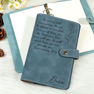 May include: A blue leather journal with a snap closure, featuring the quote "Be strong and courageous" and a floral design. The name "Dawn" is also inscribed. The journal is open, revealing blank pages.