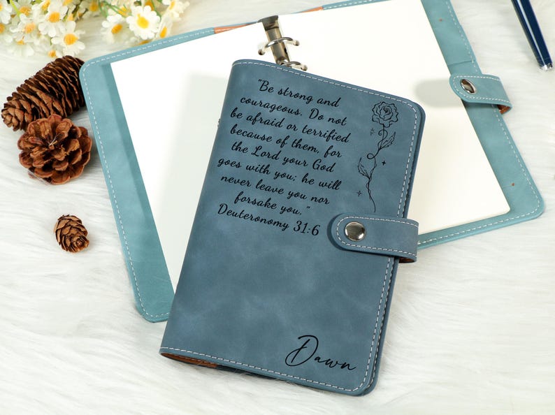May include: A blue leather journal with a snap closure, featuring the quote "Be strong and courageous" and a floral design. The name "Dawn" is also inscribed. The journal is open, revealing blank pages.
