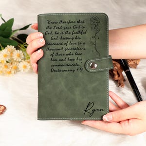 May include: A sage green leather journal with a snap closure, featuring engraved text and a floral design. The journal includes the name "Ryan" and a quote from Deuteronomy 7:9. The journal is held in a person's hands.