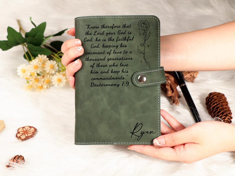 May include: A sage green leather journal with a snap closure, featuring engraved text and a floral design. The journal includes the name "Ryan" and a quote from Deuteronomy 7:9. The journal is held in a person's hands.