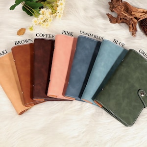 May include: A collection of leather-bound journals in various colors, including khaki, brown, coffee, pink, denim blue, blue, and green. Each journal is displayed open, showcasing the interior pages. The journals are arranged on a white, textured surface.
