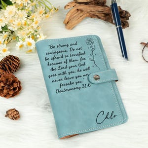 May include: A light blue leatherette journal with a snap closure. The cover features an inspirational quote and the name "Chlo" in script. A pen, glasses, and decorative items are in the background.