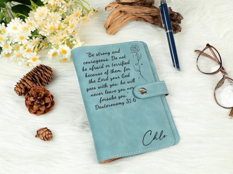 May include: A light blue leatherette journal with a snap closure. The cover features an inspirational quote and the name "Chlo" in script. A pen, glasses, and decorative items are in the background.