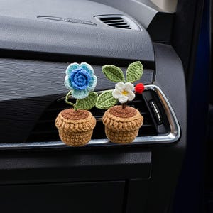 May include: Two crochet flower car air vent decorations. One has a blue flower, the other a white flower. Both have green leaves and are in brown pots. A red bead is next to the white flower.