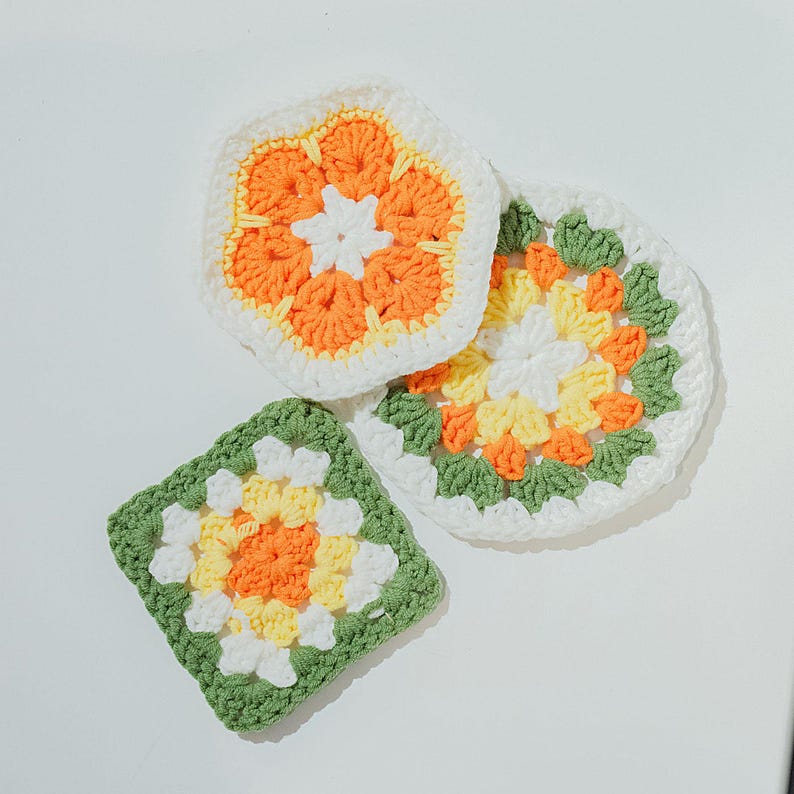 May include: A selection of handmade crochet coasters in various shapes. The coasters feature a floral design with white, orange, yellow, and green colours. The coasters are square, hexagonal, and circular.
