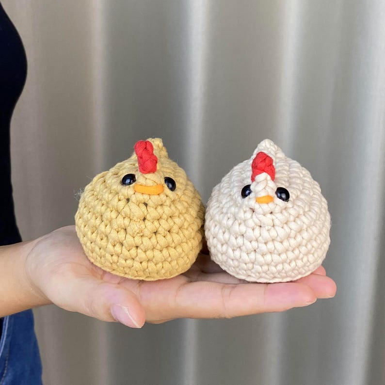 K&ouml;nnte beinhalten: Zwei handgefertigte H&auml;kelfiguren in Form von H&uuml;hnern. Eine ist hellgelb, die andere cremefarben. Beide haben rote K&auml;mme, orangefarbene Schn&auml;bel und schwarze Augen. Die H&uuml;hner sind klein, rund und haben ein strukturiertes Aussehen, was auf eine weiche, garnbasierte Konstruktion hindeutet.