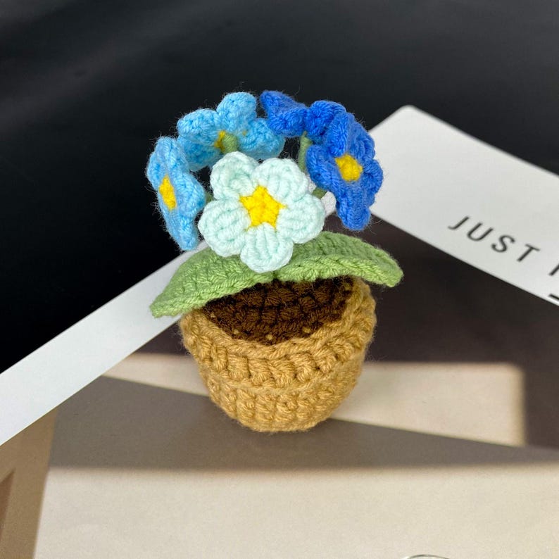 Puede incluir: Una maceta de flores hecha a mano con flores azules y celeste con centros amarillos. La maceta es marr&oacute;n y las hojas son verdes. El texto "JUST" es visible en el fondo.