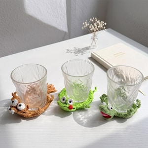 May include: Three clear, textured glass tumblers resting on individual crocheted animal coasters. One coaster is brown with a tail, one is bright green with a red mouth, and the third is light green with a pink mouth, all featuring googly eyes.