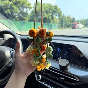 May include: A crocheted hanging ornament featuring a central tan sphere adorned with multiple small, dangling flowers in shades of yellow, orange, and cream, accented with green crocheted leaves and stems, suspended by a thin brown cord.