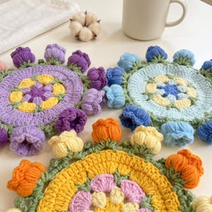Puede incluir: Tres posavasos circulares de ganchillo con un patrón floral central en amarillo, morado y rosa, rodeados por un borde de capullos de flores de ganchillo tridimensionales en tonos morado, azul, amarillo y naranja con tallos verdes.