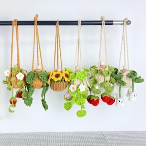 Puede incluir: Una colección de maceteros colgantes de ganchillo hechos a mano. Cada macetero presenta un diseño único, que incluye girasoles, fresas y varios acentos florales y de hojas. Los maceteros están suspendidos de una barra negra con cuerdas beige.