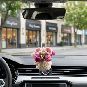 以下が含まれることがあります： ピンクと紫の花のバスケットが付いた、手作りのかぎ針編みの車の芳香剤。 花はバックミラーから吊り下げられています。 車のダッシュボードとぼやけた通りのシーンが背景に見えます。