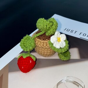 Puede incluir: Planta de fresa tejida a ganchillo con una fresa roja, hojas verdes y una flor blanca con centro amarillo. La planta est&aacute; en una maceta marr&oacute;n peque&ntilde;a. El texto "JUST FOR" es visible en el fondo.