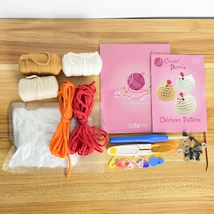K&ouml;nnte beinhalten: Ein H&auml;kelset f&uuml;r Anf&auml;nger mit Garn in Beige-, Wei&szlig;- und Cremefarben sowie orangefarbenem und rotem Garn. Das Set enth&auml;lt H&auml;kelnadeln, Maschenmarkierer und ein rosa Heft mit dem Titel "H&auml;kelanleitung f&uuml;r Anf&auml;nger" und ein Heft "Chickens Pattern".