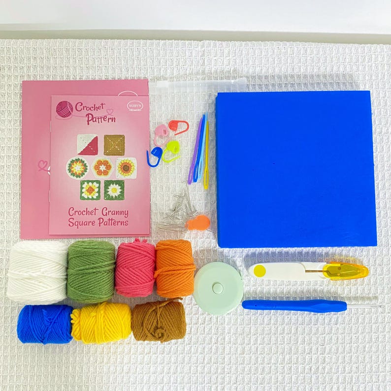 May include: A crochet kit featuring a pink booklet titled "Crochet Granny Square Patterns" and several balls of yarn in various colours. The kit includes a blue square, a measuring tape, a seam ripper, a crochet hook, and stitch markers. Yarn colours include white, green, pink, orange, blue, yellow, and brown.