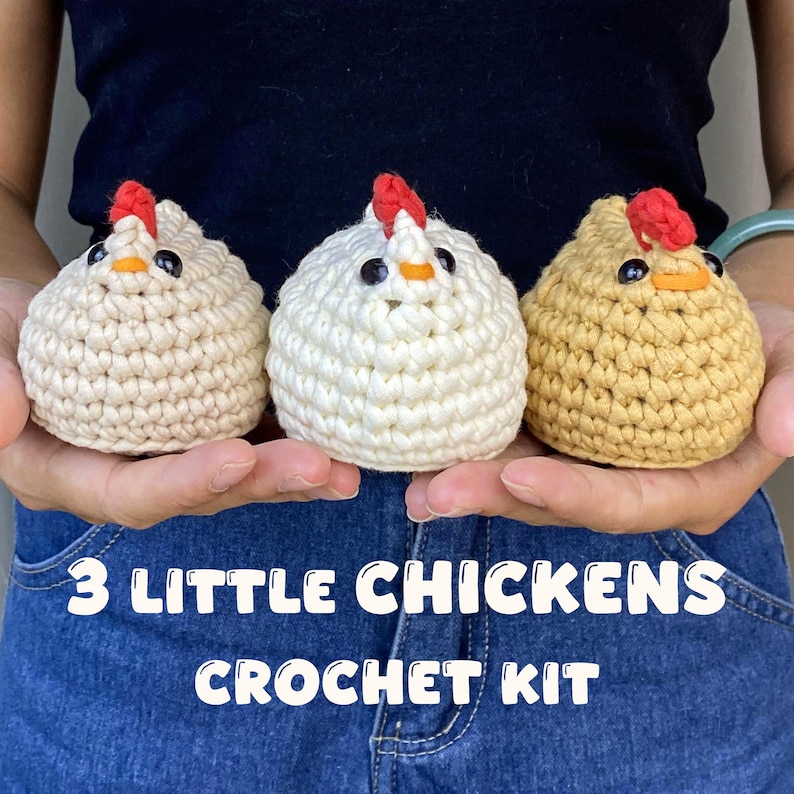 K&ouml;nnte beinhalten: Drei geh&auml;kelte H&uuml;hnerfiguren in Creme, Wei&szlig; und Gelb, jeweils mit rotem Kamm, schwarzen Augen und einem orangefarbenen Schnabel. Der Text "3 LITTLE CHICKENS CROCHET KIT" ist darunter zu sehen.