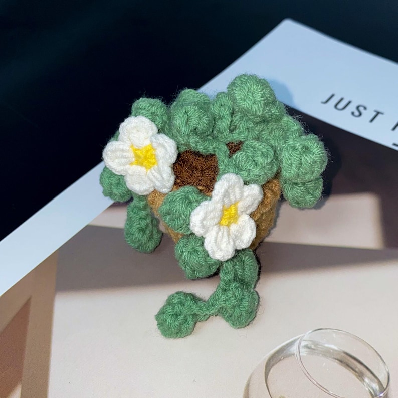 Puede incluir: Una planta de ganchillo hecha a mano en una maceta peque&ntilde;a. La planta presenta hojas verdes, flores blancas con centros amarillos y una maceta marr&oacute;n. La planta est&aacute; sobre una superficie blanca. El texto "JUST" es parcialmente visible.