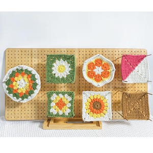 May include: A collection of colourful, handmade crochet squares in various floral and geometric designs. The squares feature shades of white, green, yellow, orange, pink, and brown. They are displayed on a wooden pegboard, highlighting the intricate details of the crochet work.