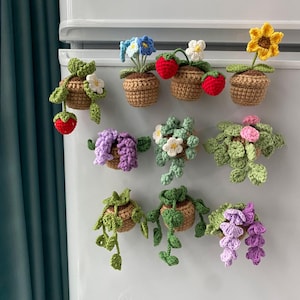 May include: A selection of handmade crochet plant magnets. The magnets depict small brown pots with colourful flowers, strawberries, and trailing vines. The flowers are in shades of yellow, blue, white, pink, and purple.