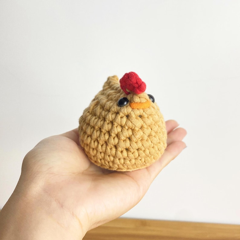 K&ouml;nnte beinhalten: Eine handgefertigte, geh&auml;kelte H&uuml;hnerfigur. Das Huhn ist hellbraun mit einem roten Kamm, einem orangefarbenen Schnabel und schwarzen Augen. Das Huhn ist klein und rund und scheint aus weichem Garn zu bestehen. Die Figur wird in einer Hand gehalten.