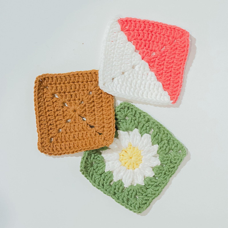 May include: Four handmade crochet squares in different colours. One is brown, another is white and coral, and the third is green with a white and yellow daisy. Ideal for crafting projects.