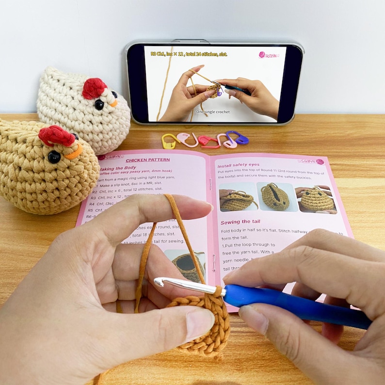 K&ouml;nnte beinhalten: Ein H&auml;kelprojekt mit einem H&uuml;hnermuster, Garn und einem H&auml;kelhaken. Das Bild zeigt ein Smartphone mit einem H&auml;keltutorial, geh&auml;kelten H&uuml;hnerspielzeugen und einem Musterbuch mit Anleitungen.
