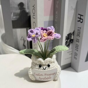 May include: A crocheted potted plant featuring light purple and lavender flowers with yellow centers, green leaves, and a white crocheted pot with a smiling face. A white sign attached to the front reads Custom Message Available.