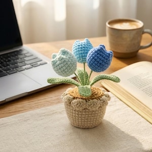 May include: A crochet potted plant featuring three tulip-shaped flowers in shades of light blue, medium blue, and dark blue. The flowers have green crocheted leaves and are set in a beige crocheted pot with a textured, scalloped top edge.