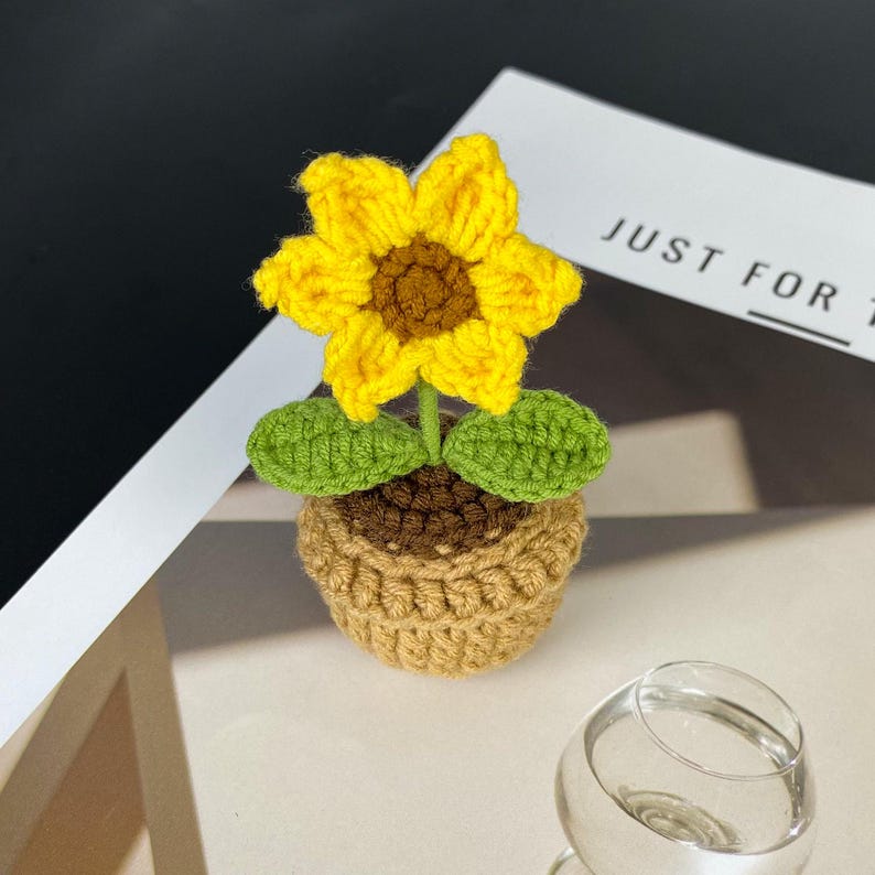 Puede incluir: Un girasol de ganchillo hecho a mano en una maceta peque&ntilde;a. El girasol tiene p&eacute;talos de color amarillo brillante y un centro marr&oacute;n. Hojas verdes y una maceta marr&oacute;n completan el dise&ntilde;o. La maceta es de color marr&oacute;n claro y el texto "JUST FOR" es visible en el fondo.