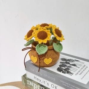Puede incluir: Arreglo de girasoles de ganchillo en una cesta marrón con detalles de corazón amarillo. Los girasoles tienen pétalos amarillos y centros marrones, con hojas verdes. La cesta está atada con una cuerda marrón. Sobre una pila de libros.