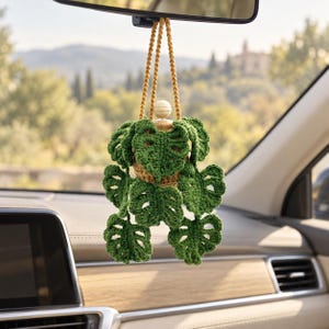May include: A crocheted monstera plant hanging ornament with dark green leaves and a brown pot, suspended by a mustard yellow cord. The plant features a small wooden bead at the top and is designed to hang from a vehicle rearview mirror.