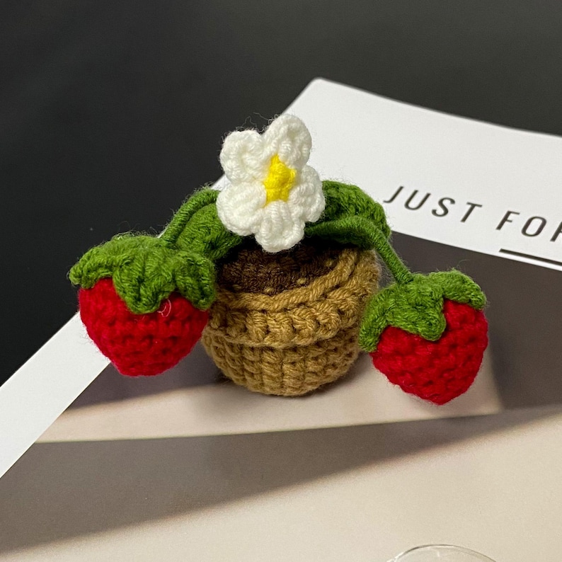 Puede incluir: Una planta de fresa de ganchillo con dos fresas rojas, hojas verdes, una flor blanca con centro amarillo y una maceta marr&oacute;n y beige. El texto "JUST FOR" es visible en el fondo.