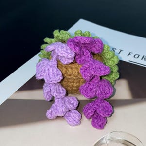 Puede incluir: Una pinza para el pelo hecha a mano con ganchillo con un racimo de flores moradas y lavanda con hojas verdes. Las flores est&aacute;n unidas a una base marr&oacute;n. La pinza est&aacute; sobre una superficie blanca con el texto "ST FOR".