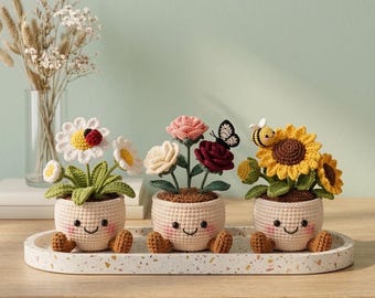 Puede incluir: Tres plantas en maceta de ganchillo con caras sonrientes sobre una bandeja moteada. La maceta izquierda tiene margaritas blancas y una mariquita, la central tiene rosas rosas y rojo oscuro con una mariposa, y la derecha tiene girasoles amarillos y una pequeña abeja.