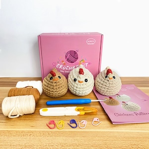 K&ouml;nnte beinhalten: Ein rosa H&auml;kel-Set mit dem Text "CROCHET KIT" und drei geh&auml;kelten H&uuml;hnerfiguren. Das Set enth&auml;lt Garn, eine H&auml;kelnadel, Maschenmarkierer und ein Musterheft. Die H&uuml;hner sind beige, braun und wei&szlig;.