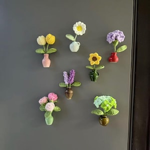 May include: A selection of handmade crochet flower magnets in various colours, including yellow, white, purple, pink, and green. Each flower is in a small ceramic vase. The magnets are arranged on a grey surface.